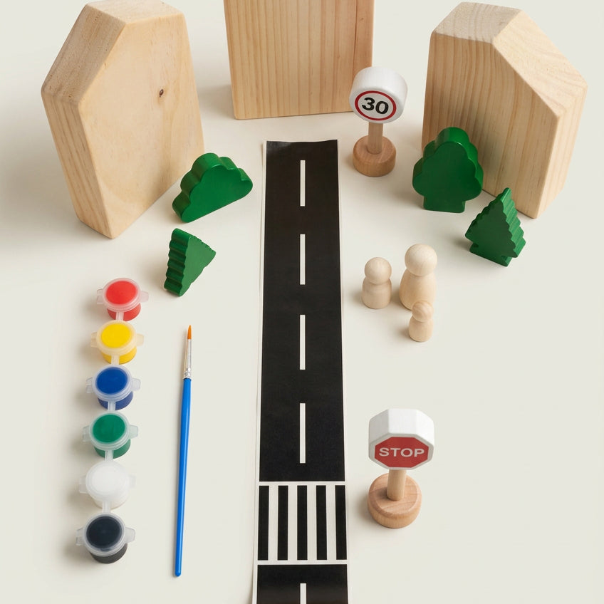 A children's wooden toy road playset arranged on a light beige surface, including a black road with white dashed lines, wooden house shapes, green trees, small wooden cars, a paint palette with a blue paintbrush, and a speed-limit sign.