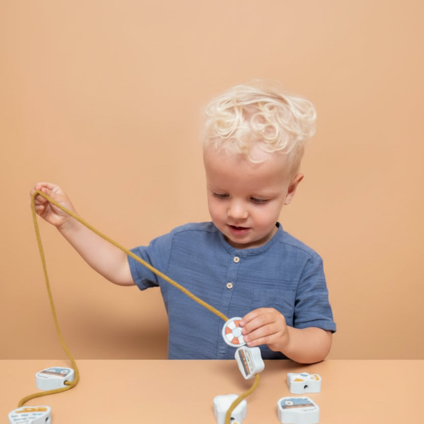 A young child with blonde hair wearing a blue shirt is sitting at a table playing with a beading set, and reel while several small fish-shaped pieces are scattered on the table against a solid beige background.