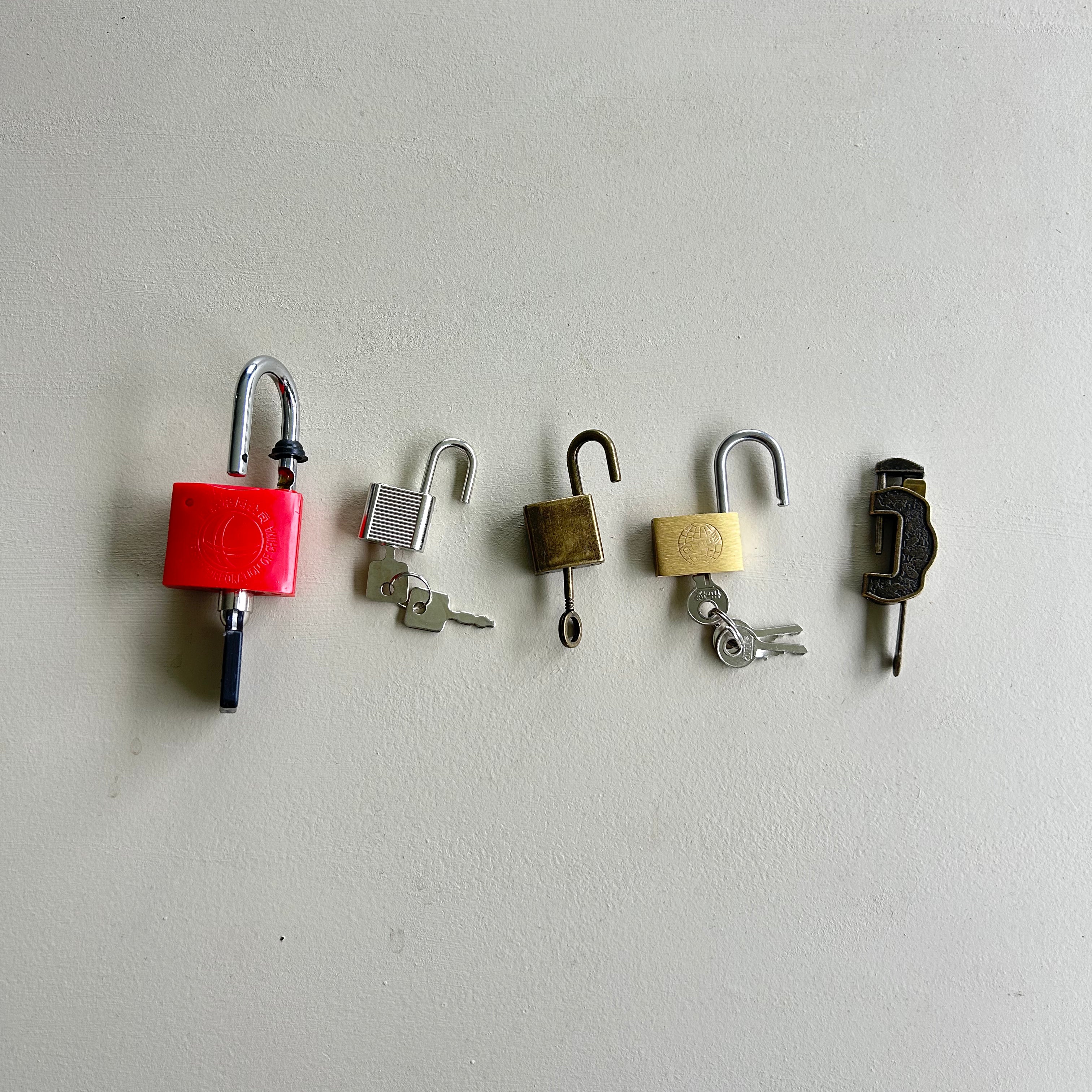Five different padlocks and keys arranged in a row against a plain off-white wall. The locks include a bright red cylindrical padlock, a silver metal padlock with a key, a small bronze padlock, a cream padlock with a key, and a dark metal padlock. montessori