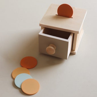 A minimalist wooden drawer toy with a partially open drawer and a small orange circular piece on top, shown on a warm off-white background with three loose matching pieces in orange, yellow, and light blue.