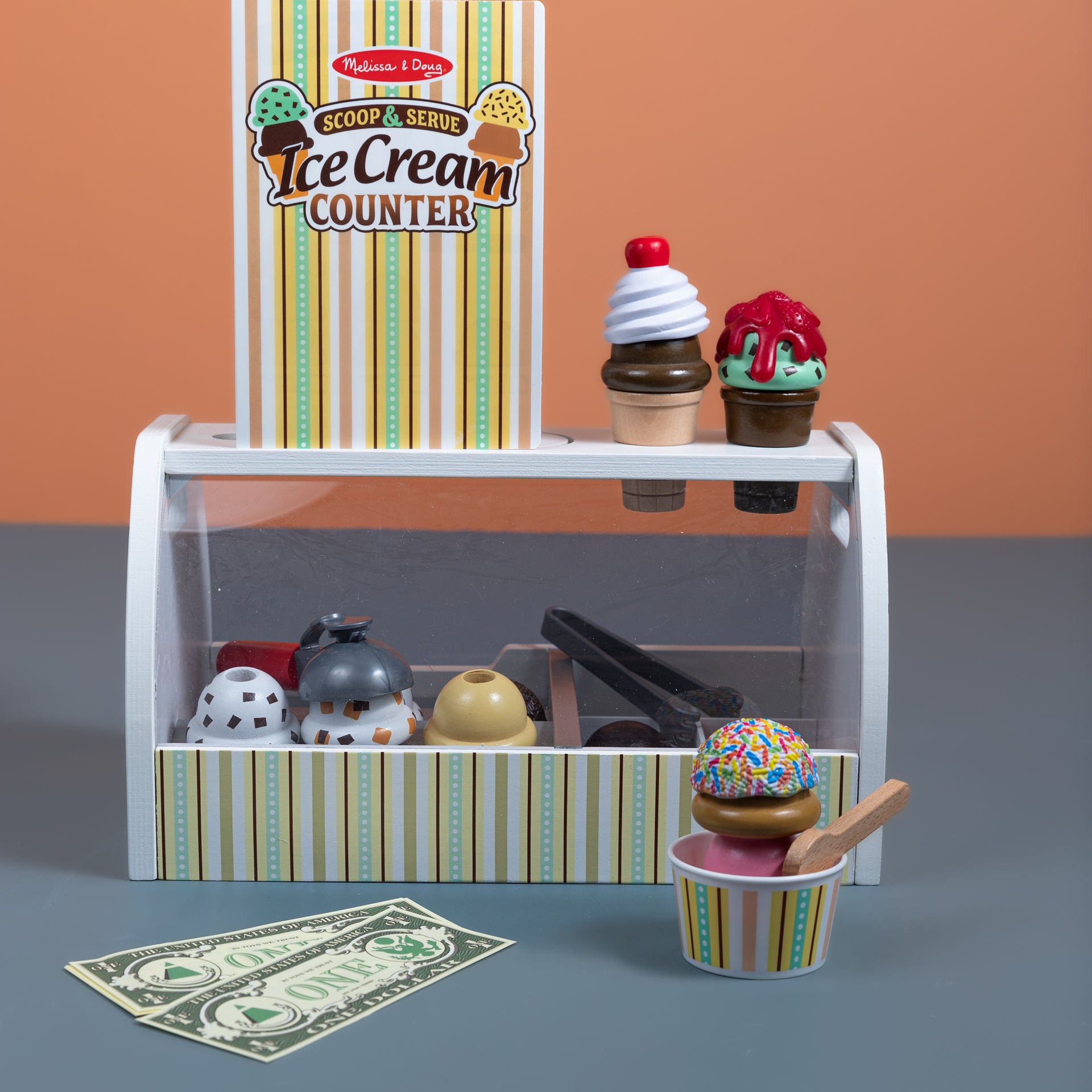 Melissa & Doug Scoop & Serve Ice Cream Counter wooden playset displayed with all 28 pieces including scoops, toppings, cones, cup, scooper, tongs, spoon, menu card, and pretend dollar bills arranged in front of the wooden counter