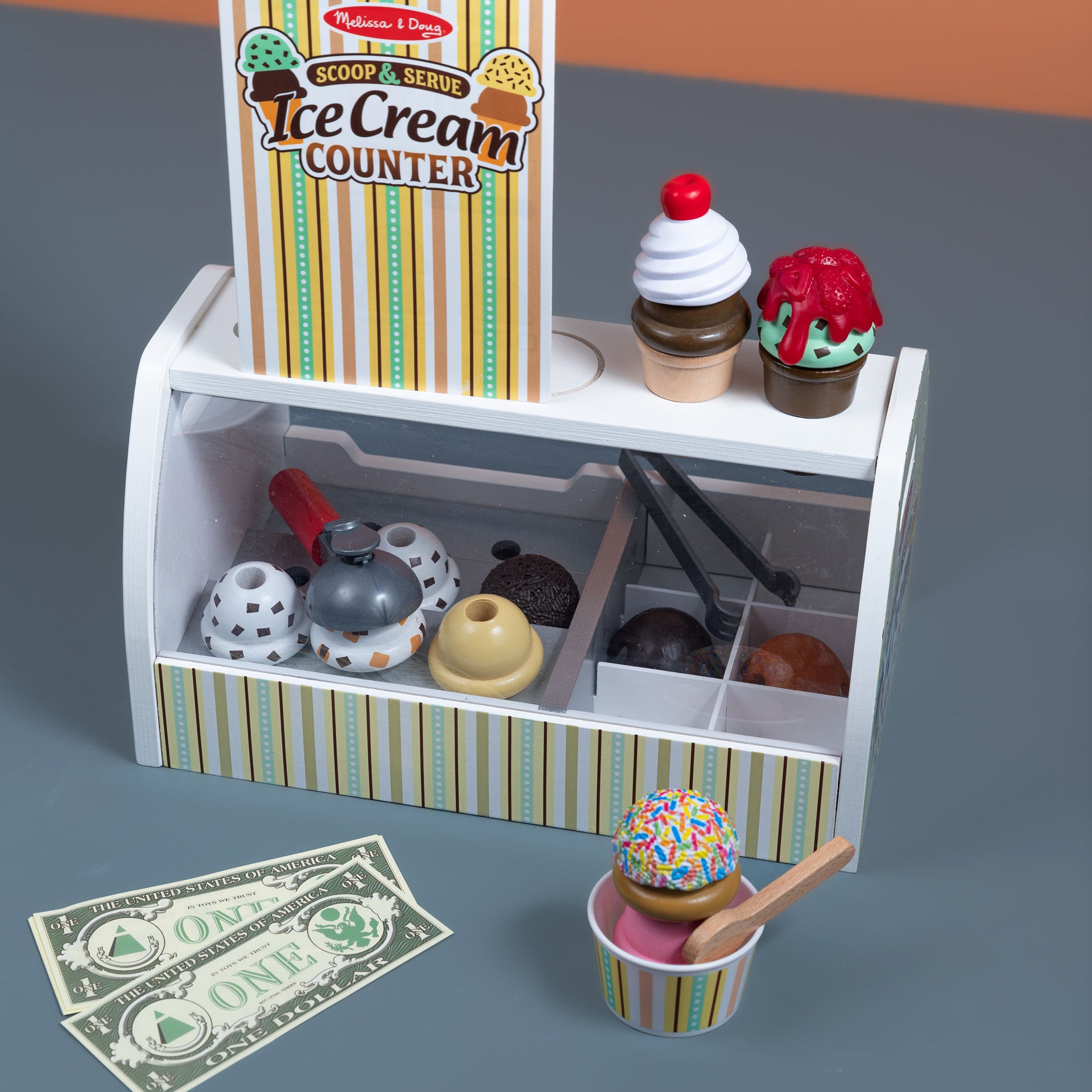 Toy ice cream counter set with ice cream cones, toppings, and money on a gray surface.