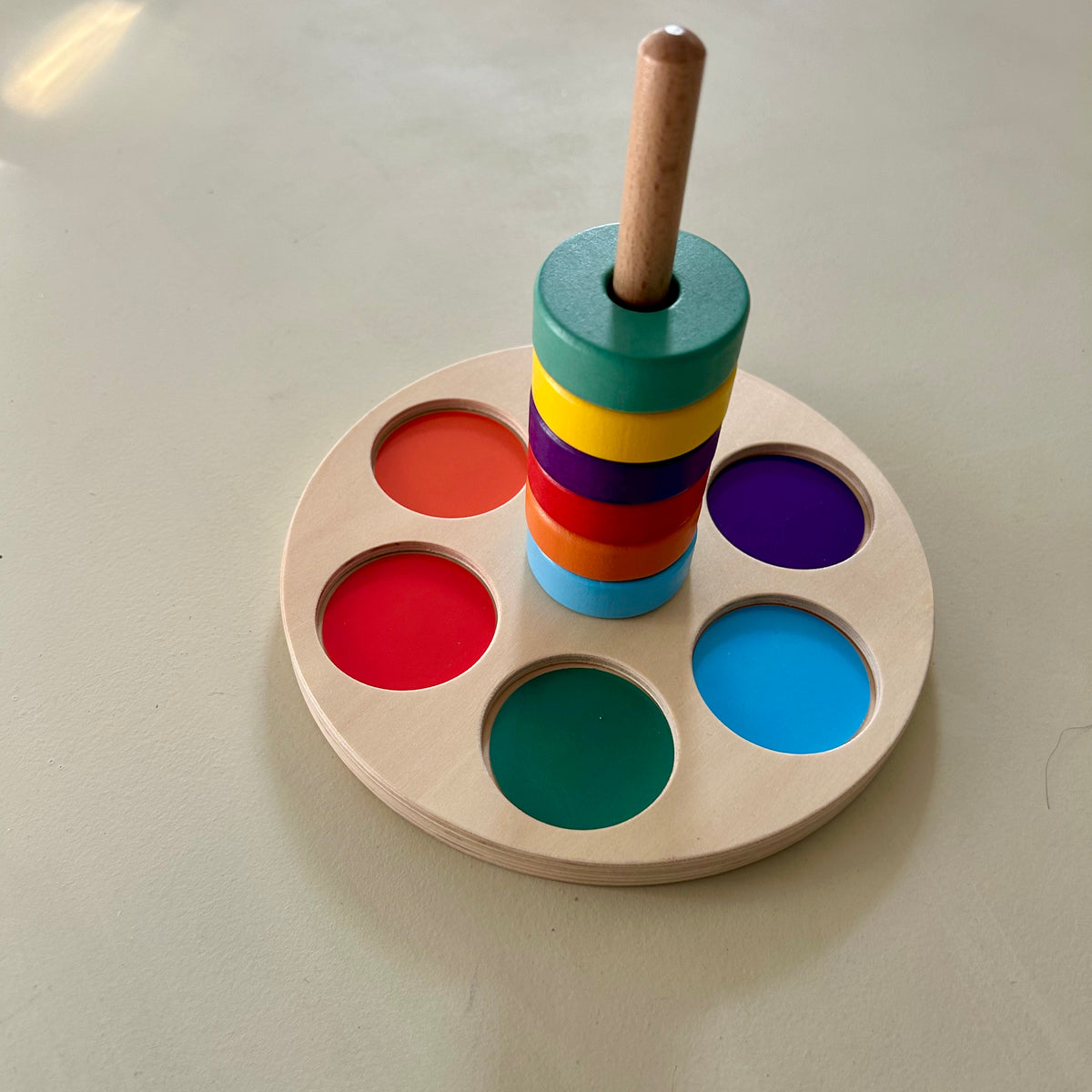 Wooden colour matching stacker on a pale sage surface. Six painted wooden rings are stacked on a central wooden peg: light blue, orange, red, purple, yellow and green. They sit on a round natural plywood base with recessed circles in orange, red, green, light blue and purple.
