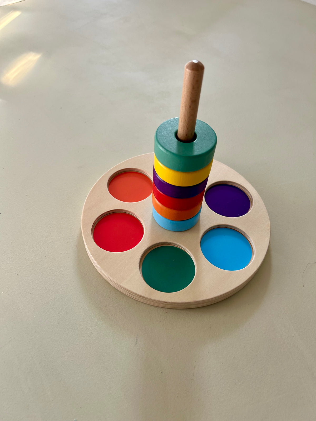 Wooden colour matching stacker on a pale sage surface. Six painted wooden rings are stacked on a central wooden peg: light blue, orange, red, purple, yellow and green. They sit on a round natural plywood base with recessed circles in orange, red, green, light blue and purple.