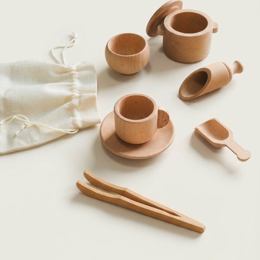 A collection of wooden sensory play tools including a pot with lid, cup, plate dismeter, bowl, scoops, tweezer, and two wooden sticks, displayed on a green surface with a drawstring bag.