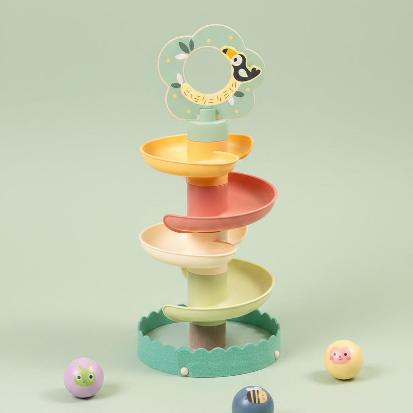A pastel spiral toy with four winding levels in mint green, yellow, peach, and cream, topped with a flower-shaped ring holder. Small painted balls are placed around it on a light beige textured surface against a soft cream background.