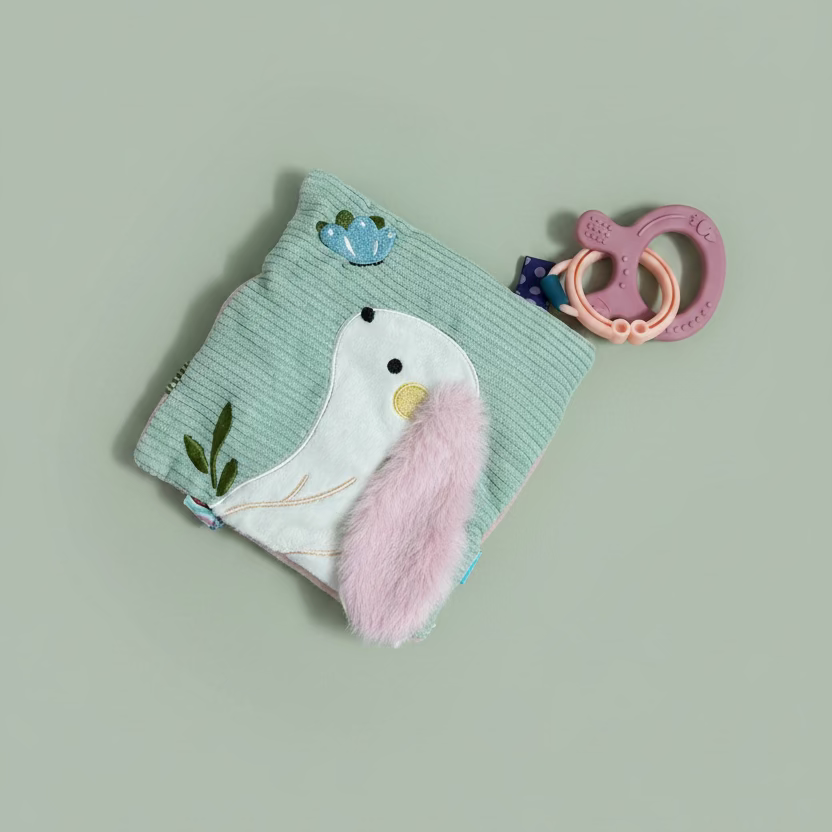A baby teething toy set on a light wood surface, featuring a mint green corduroy pouch with a white bird applique, a fluffy pink ear, and a pink silicone teether ring.