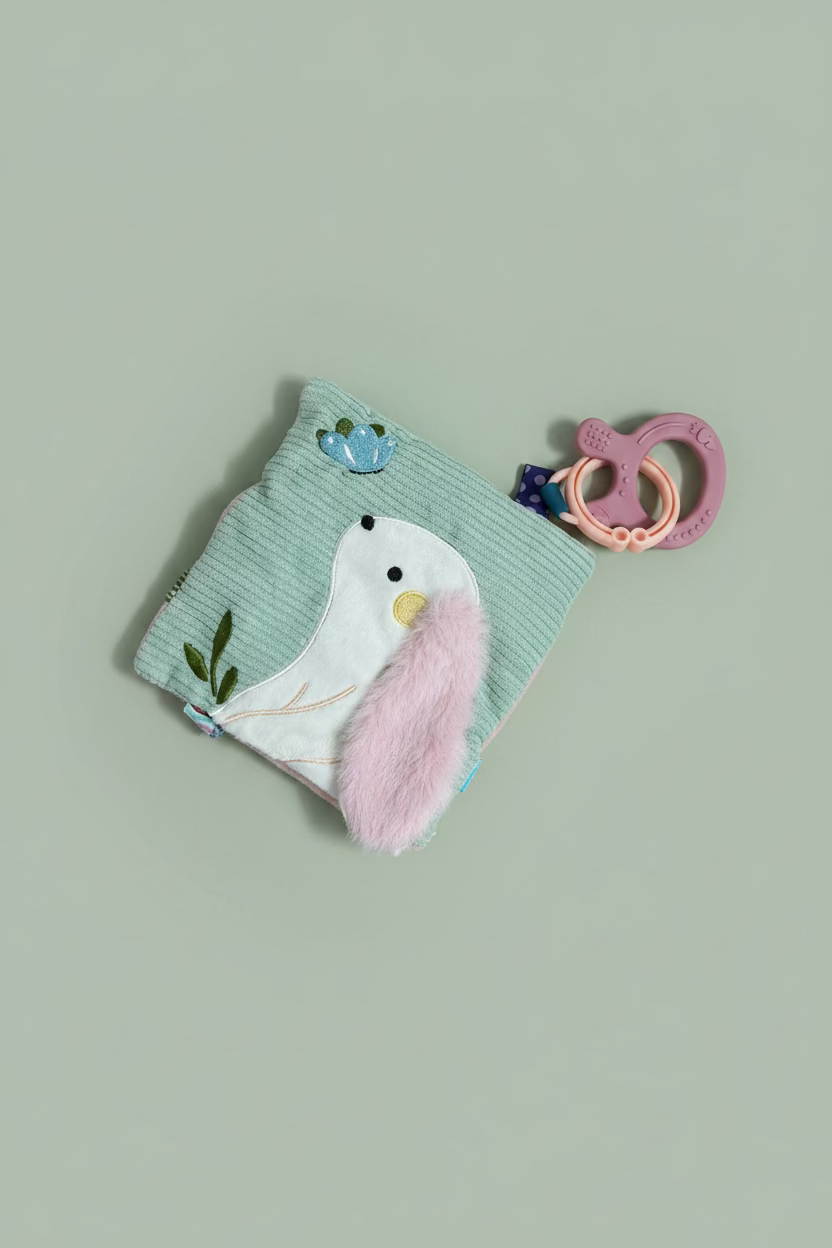 A baby teething toy set on a light wood surface, featuring a mint green corduroy pouch with a white bird applique, a fluffy pink ear, and a pink silicone teether ring.