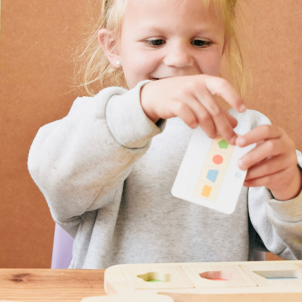 A child’s hands turning a wooden shape block to fit it onto the whale board, with a puzzle card propped up beside them.