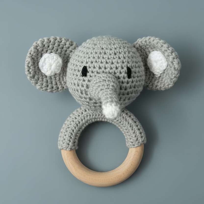 A handmade crocheted elephant rattle with a natural wooden ring handle, resting on a light-colored wooden surface.