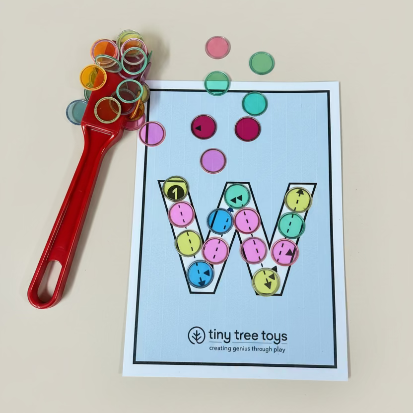 A children's letter learning game card featuring the letter 'W' filled with colorful buttons, shown on a light wood tabletop next to a red spoon holding more colorful buttons. The brand 'tiny tree toys' is printed at the bottom of the card.