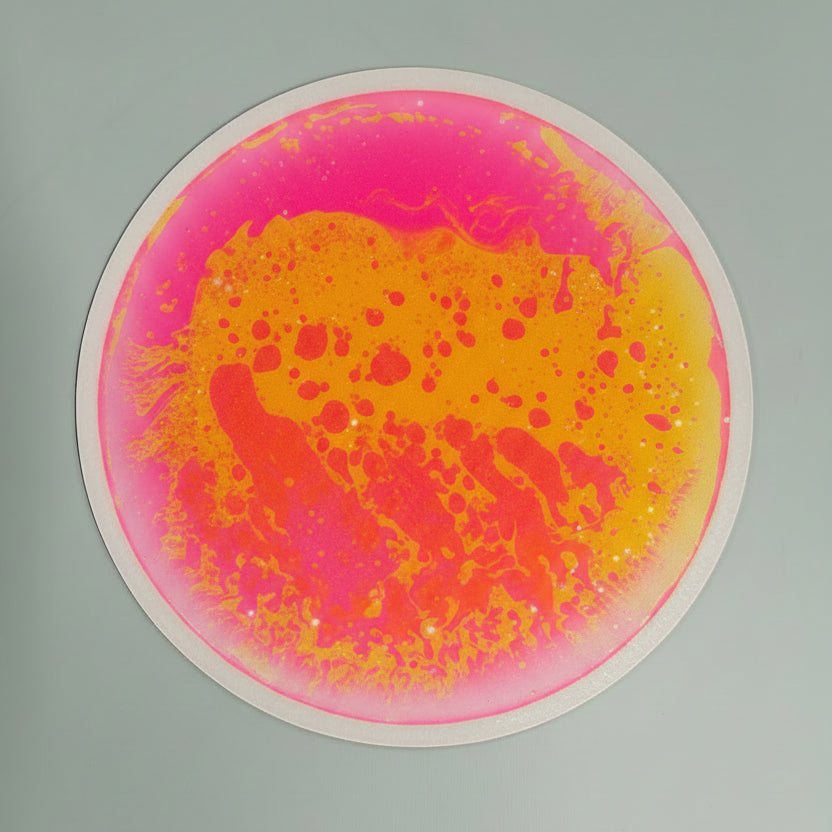 A circular liquid tile with a vibrant abstract design featuring bright pink and yellow-orange paint splatters on a light gray background.