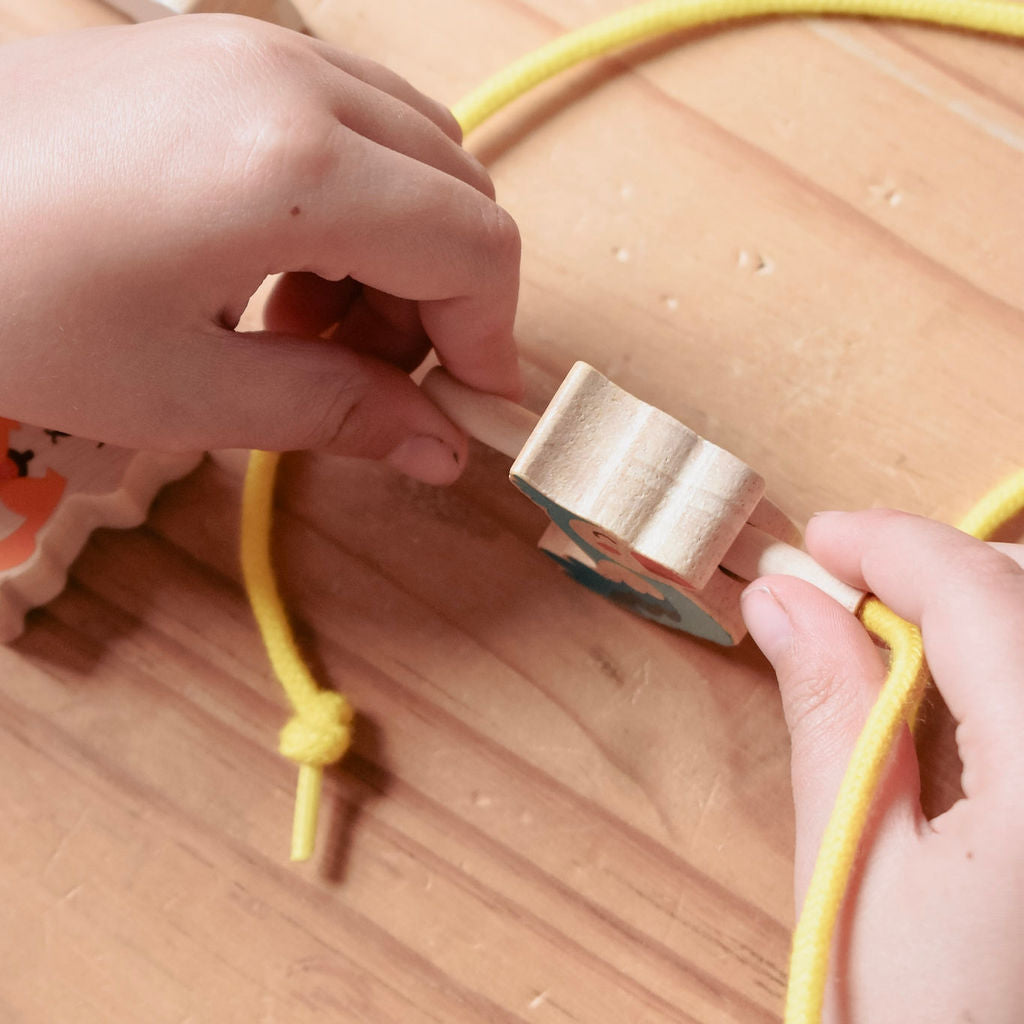 A close-up of hands tying a yellow rope around a small wooden bead on a light wood surface.