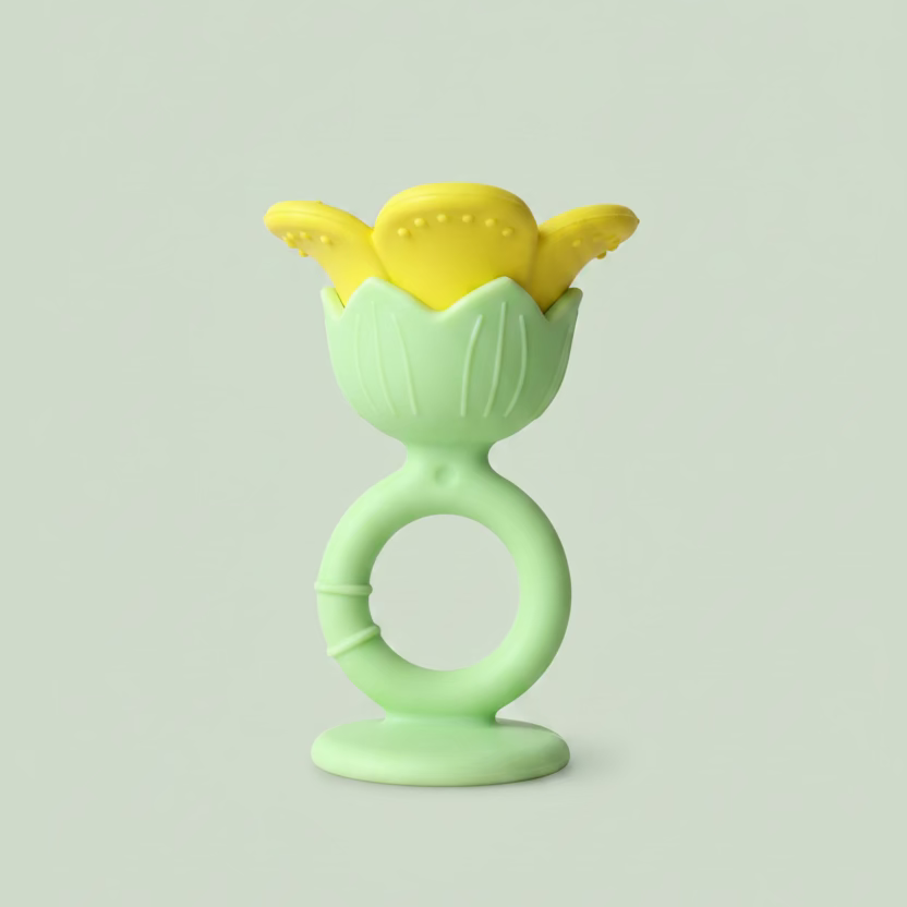 A baby teether toy with a light green silicone ring handle and a yellow flower-shaped silicone nibbler on top, resting on a light-colored wooden surface.