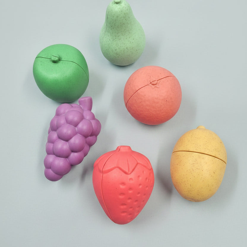 Six pastel fruit-shaped items arranged on a warm peach background: a green apple, a mint pear, a pink grapefruit, a purple grape cluster, a pink strawberry, and a pale yellow lemon. Each item has a split seam across the top half, suggesting they may be capsules, containers, or novelty fruit pieces.