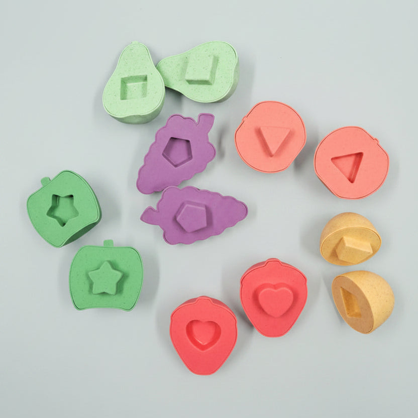 A set of colorful wooden shape-sorting blocks and matching cutters arranged on a soft peach background. The pieces are in pastel shades of green, pink, purple, and yellow, with different geometric and fruit-like shapes.