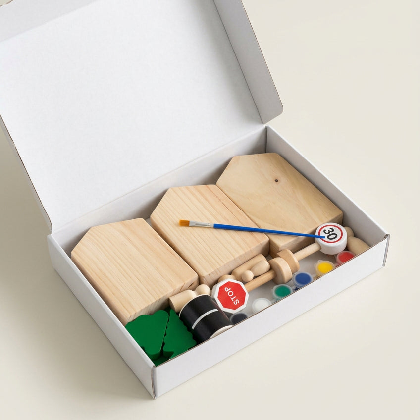 A wooden toy construction set inside an open white box, featuring light wood blocks, a small brush, colorful paint pots, and green tree pieces on a beige surface.