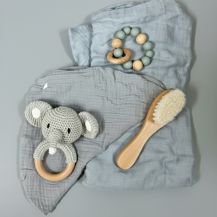 A baby gift set featuring a dusty blue muslin blanket set with a crocheted elephant teether, a wooden bead rattle, and a wooden baby brush, arranged on a light wood background.