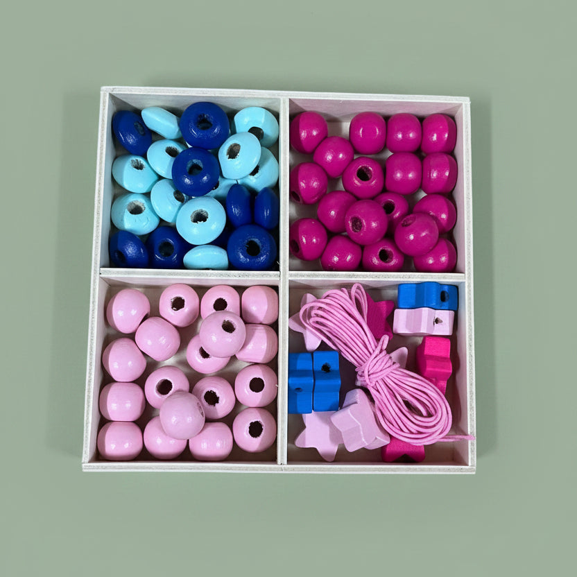 A 4-compartment tray filled with colorful wooden beads and small wooden blocks, placed on a light wood surface. The beads are in pink, blue, and magenta, with some larger round beads and some small square blocks.