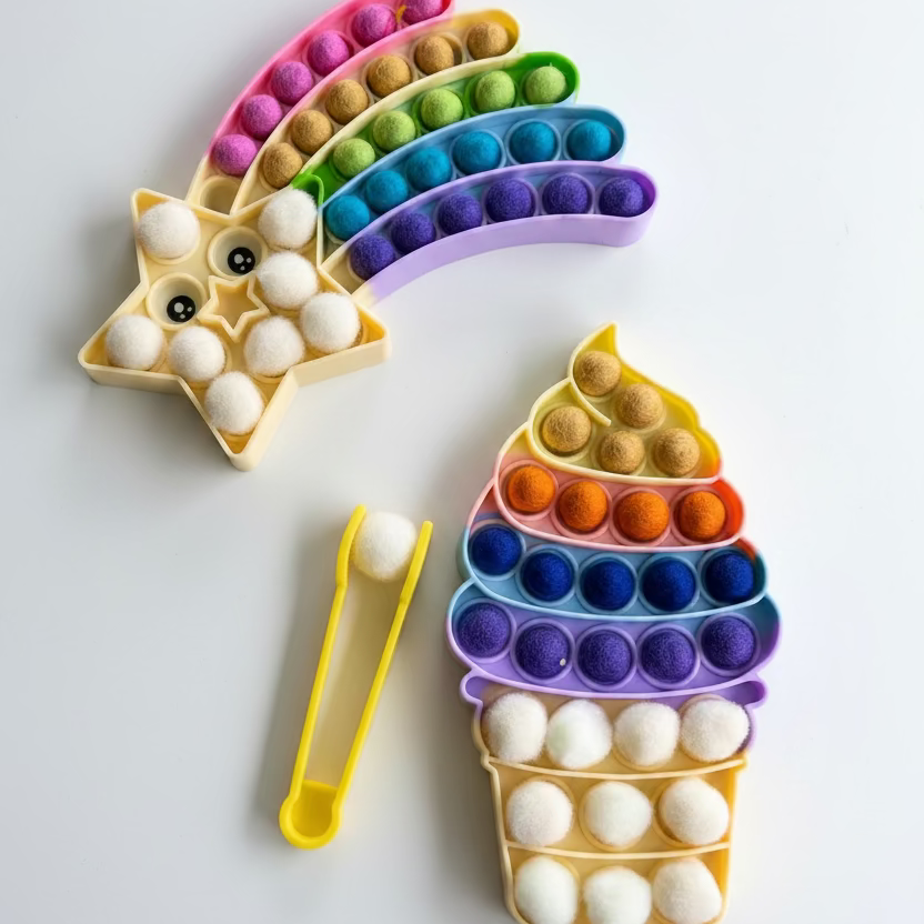 A colorful sensory toy set on a muted blue-gray background, featuring a rainbow-shaped pop-it toy, a star-shaped pop-it toy with googly eyes, a cupcake-shaped pop-it toy, and a pair of yellow tongs.