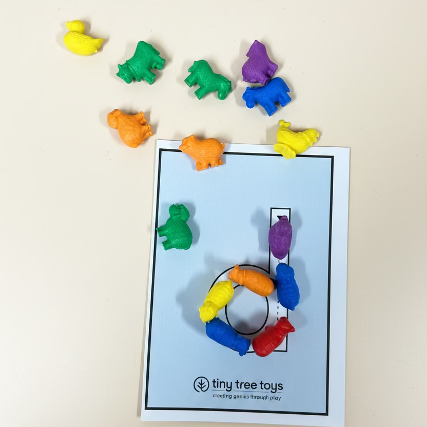 A set of small, colorful animal-shaped toy pieces arranged on a wooden surface around a printed card. The cards are white with black outlines and the brand name 'tiny tree toys' printed at the bottom.