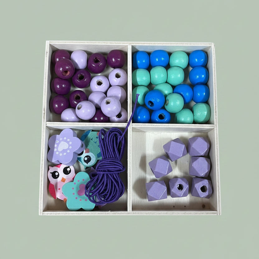 A 4-compartment wooden box filled with colorful wooden beads and small wooden charms, placed on a light wood tabletop. The beads come in purple, blue, mint green, and lavender colors.