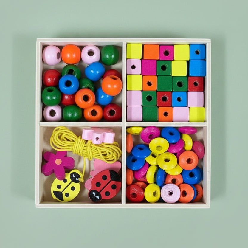 A wooden compartment box filled with colorful wooden beads and toy pieces, including round discs, square blocks, yellow cord, and ladybug-shaped beads, photographed on a light wood tabletop.