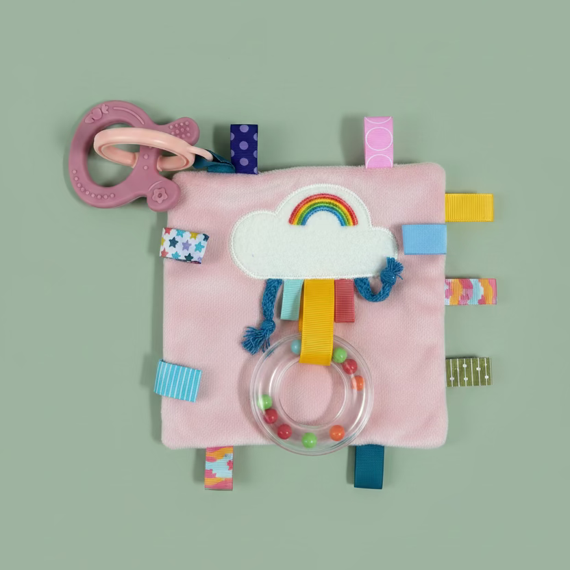 A pink textured baby sensory activity book with a cloud applique, colorful fabric tabs, and a clear plastic rattle, laid flat on a light wood surface.
