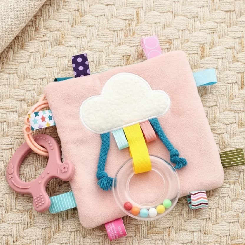 Soft square taggie comforter blanket in green and pink with a rainbow appliqué, colourful fabric tags around the edges, shown with a small ring with tiny balls rattle