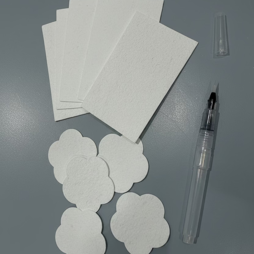 White cloud-shaped sponge shaped paper for painting arranged next to a clear water brush and a small clear cap on a light gray surface.