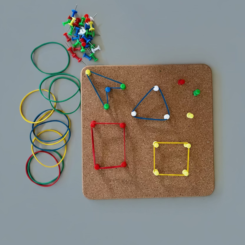 A children's geometric pegboard activity toy on a light gray background, featuring a square cork board with colorful wooden pegs, sticks, and shape frames, alongside a pile of multicolored rubber bands and loose pins.