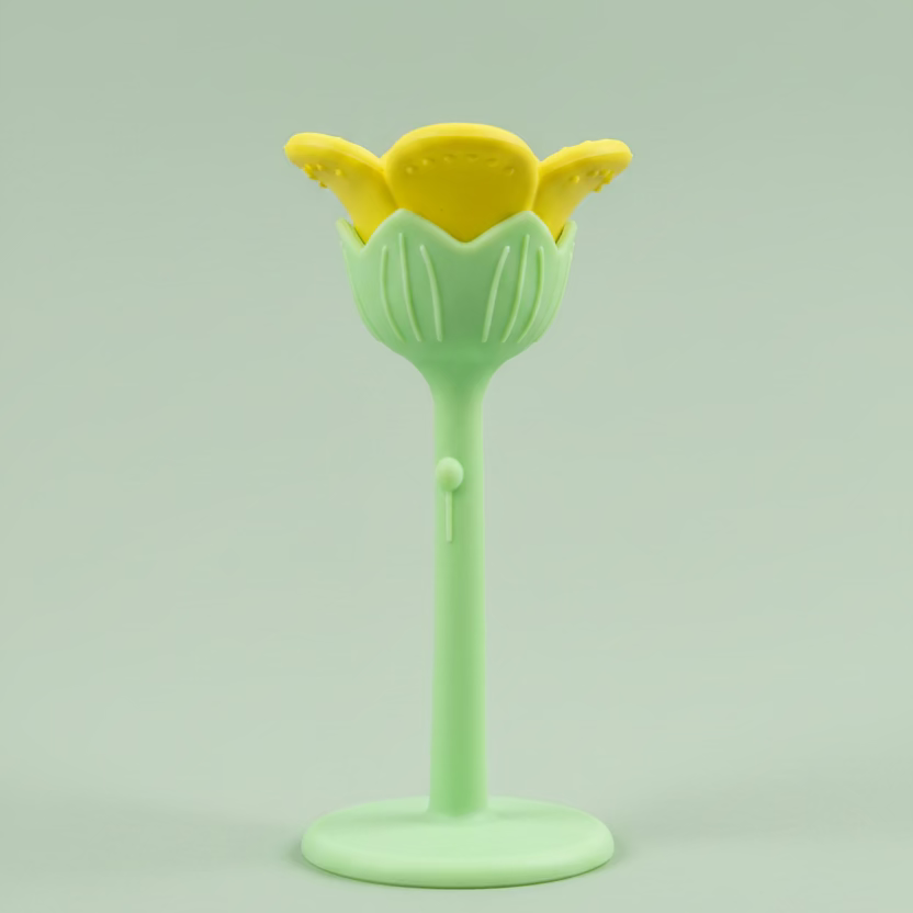 A small flower-silicone teether with a yellow top and a light green stem and base, photographed against a wooden surface.