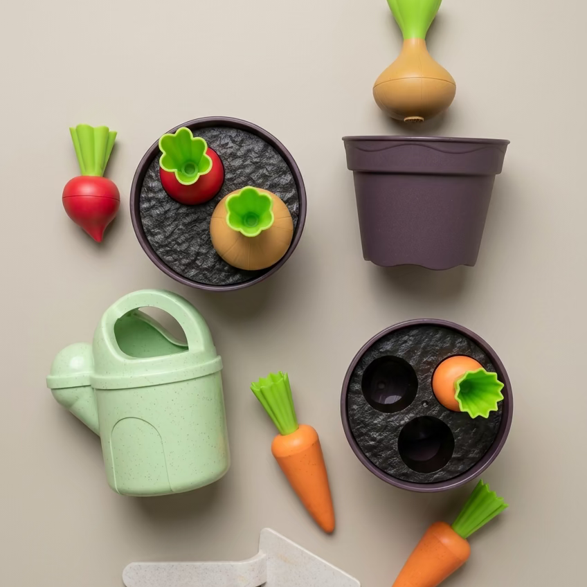 A set of wooden toy gardening items including two pots with planters, a watering can, a trowel, carrots, radishes, and an onion, arranged on a light wood background.