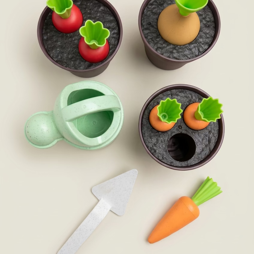 A children's wooden pretend-gardening set arranged on a light wood tabletop, including dark plant pots with toy vegetables, a mint green watering can, an orange carrot, and a silver-colored trowel.