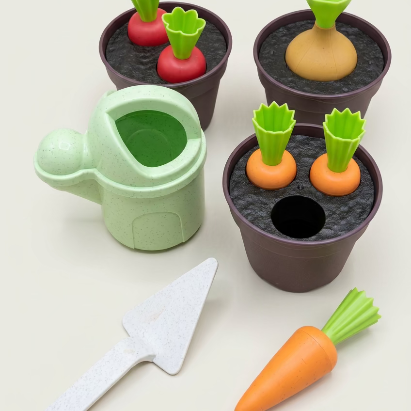 A children's toy gardening set on a wooden tabletop, including a light green watering can, dark brown pots with toy soil, orange carrots with green tops, red radishes, and a white trowel.