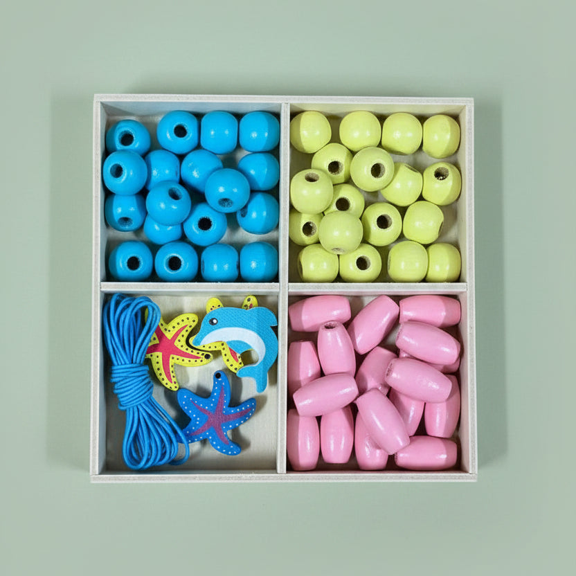 A wooden four-compartment box containing colorful craft beads: blue round beads, yellow oval beads, pink teardrop beads, and blue starfish and shark-shaped beads, placed on a wooden surface.