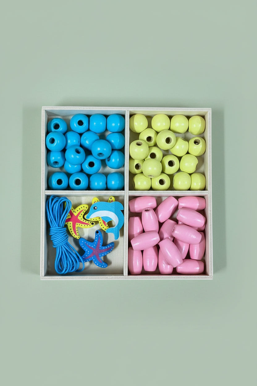 A wooden four-compartment box containing colorful craft beads: blue round beads, yellow oval beads, pink teardrop beads, and blue starfish and shark-shaped beads, placed on a wooden surface.