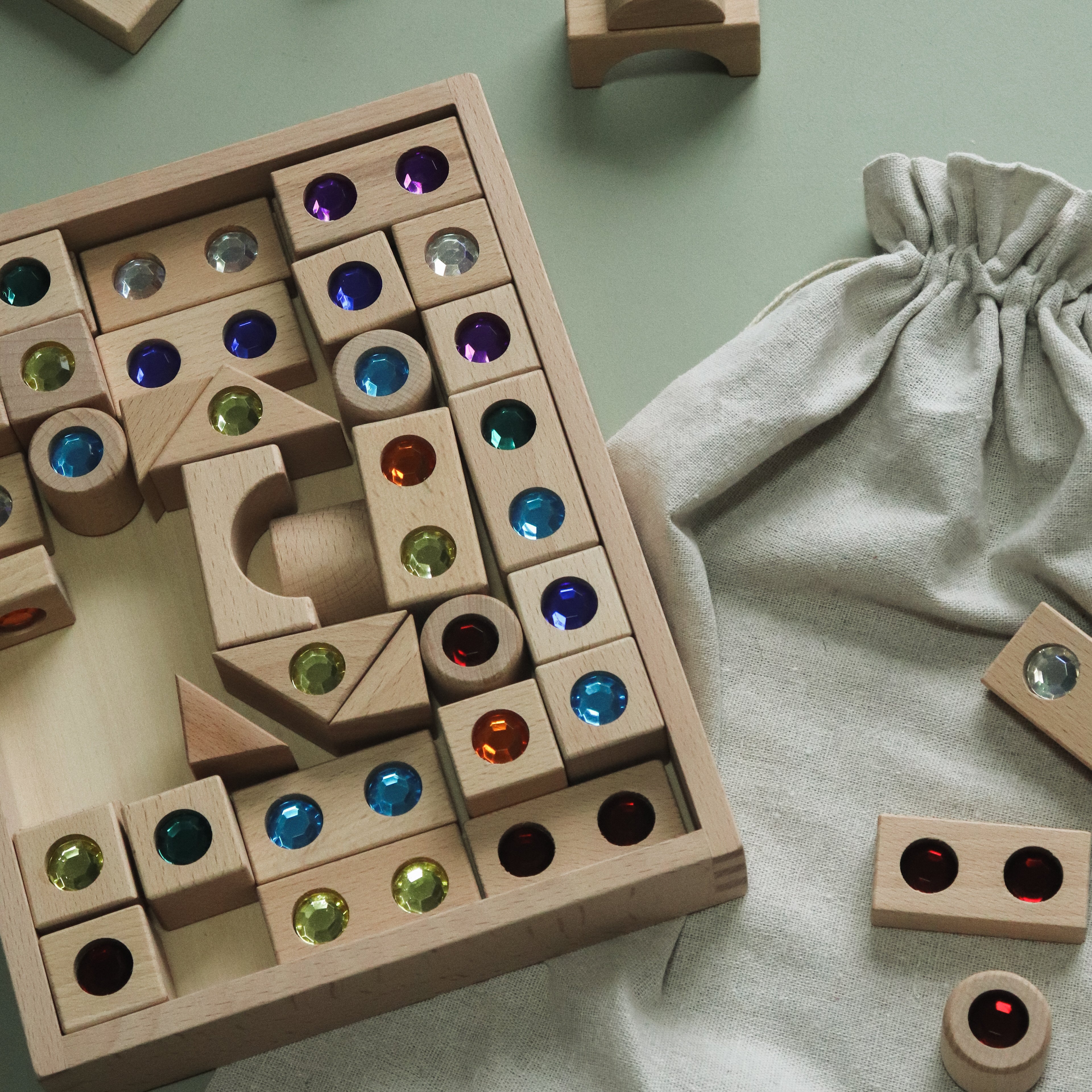 A wooden block puzzle game with colorful gemstone inserts, shown with a light gray fabric drawstring bag on a pale green background.