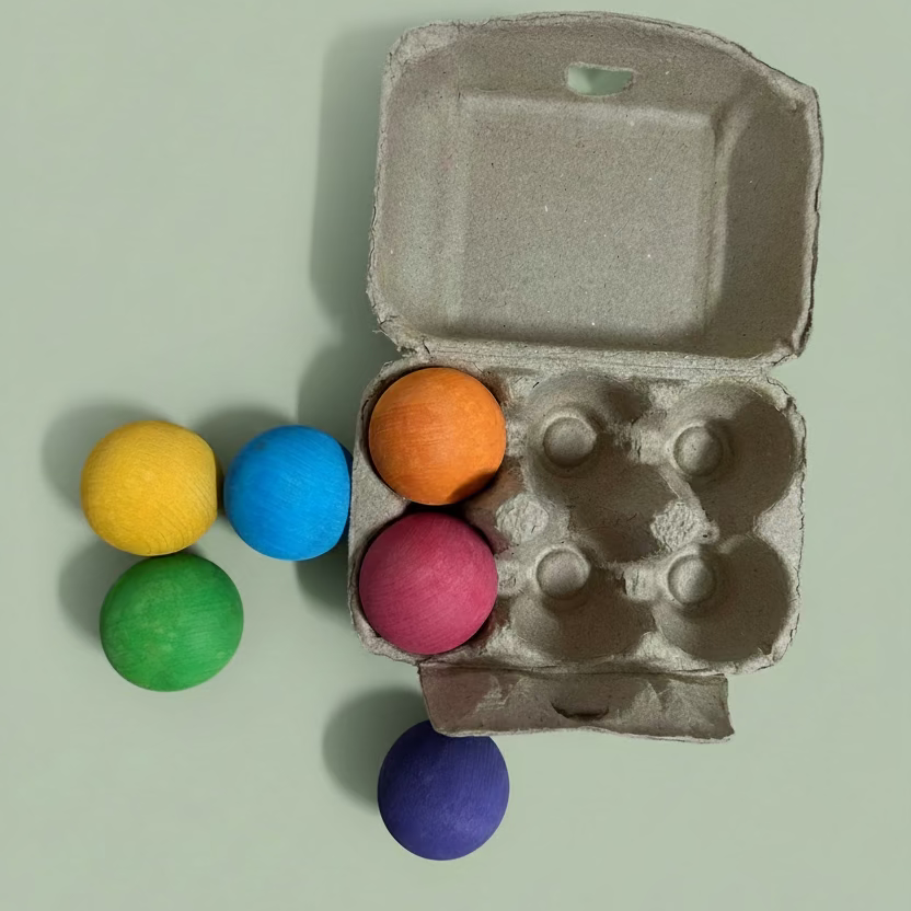A cardboard egg carton with six colorful craft wooden balls in orange, pink, blue, green, yellow, and purple.