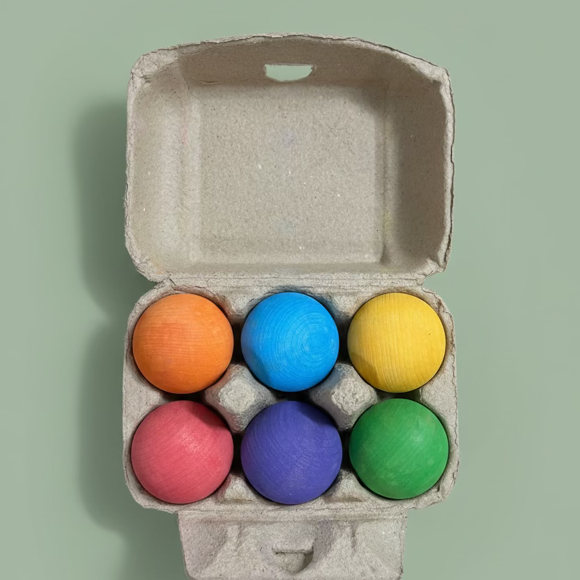 Six hand-painted wooden balls in orange, blue, yellow, pink, purple, and green, arranged inside an open brown cardboard egg carton on a light wood tabletop.