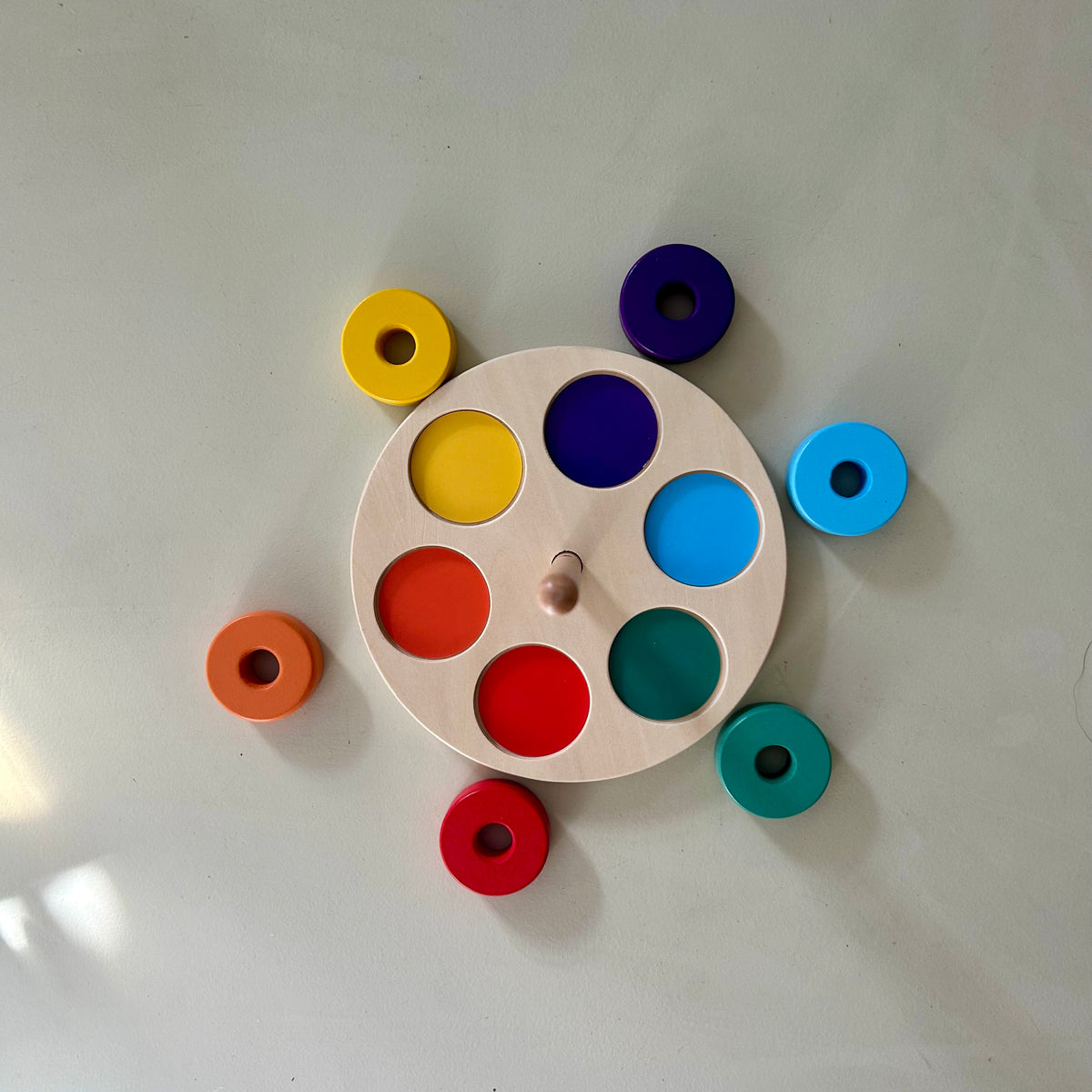 Overhead view of the round wooden base with six coloured circles, and six painted wooden rings laid out beside it in green, yellow, purple, red, orange and light blue.