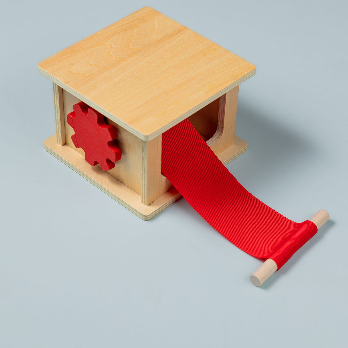 Natural wooden Montessori pulling and winding box with a red cog-shaped knob on the side and a red ribbon pulled out to a wooden dowel handle, on a pale blue-grey background.
