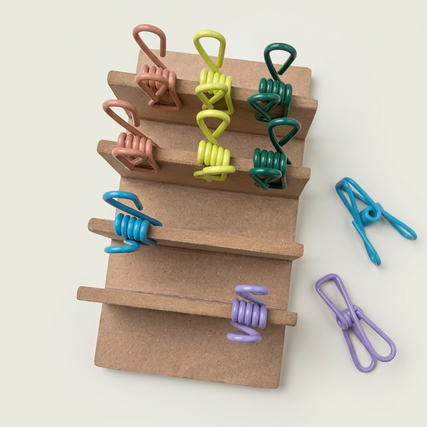A set of colorful metal spring clips in multiple colors, including blue, purple, green, yellow, and pink, displayed on a small wooden stand with a few loose clips on a light wood tabletop.