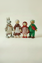 Four bendable fabric dolls standing in a row,  a grey wolf in a floral dress and apron, Little Red Riding Hood in a pink dress with red cape and hood, a grey-haired Grandmother in a floral dress and apron, and a Woodsman in a green outfit with a striped cap and axe, shown against a white background