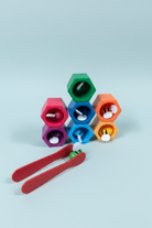 A colorful bee-themed toy set on a wooden tabletop, featuring six hexagonal bins in green, orange, blue, pink, and purple, each containing a small bee figure, with red tongs holding a green bee in the foreground.