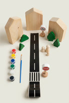 A children's wooden toy road playset arranged on a light beige surface, including a black road with white dashed lines, wooden house shapes, green trees, small wooden cars, a paint palette with a blue paintbrush, and a speed-limit sign.