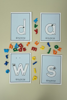 Four printable tracing cards for lowercase letters d, a, w, and s arranged on a wooden surface, surrounded by colorful magnetic alphabet letters. The brand name 'tiny tree toys' is visible at the bottom of each card.