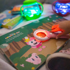 Child's hand holding a light-up toy over a children's book with colorful background