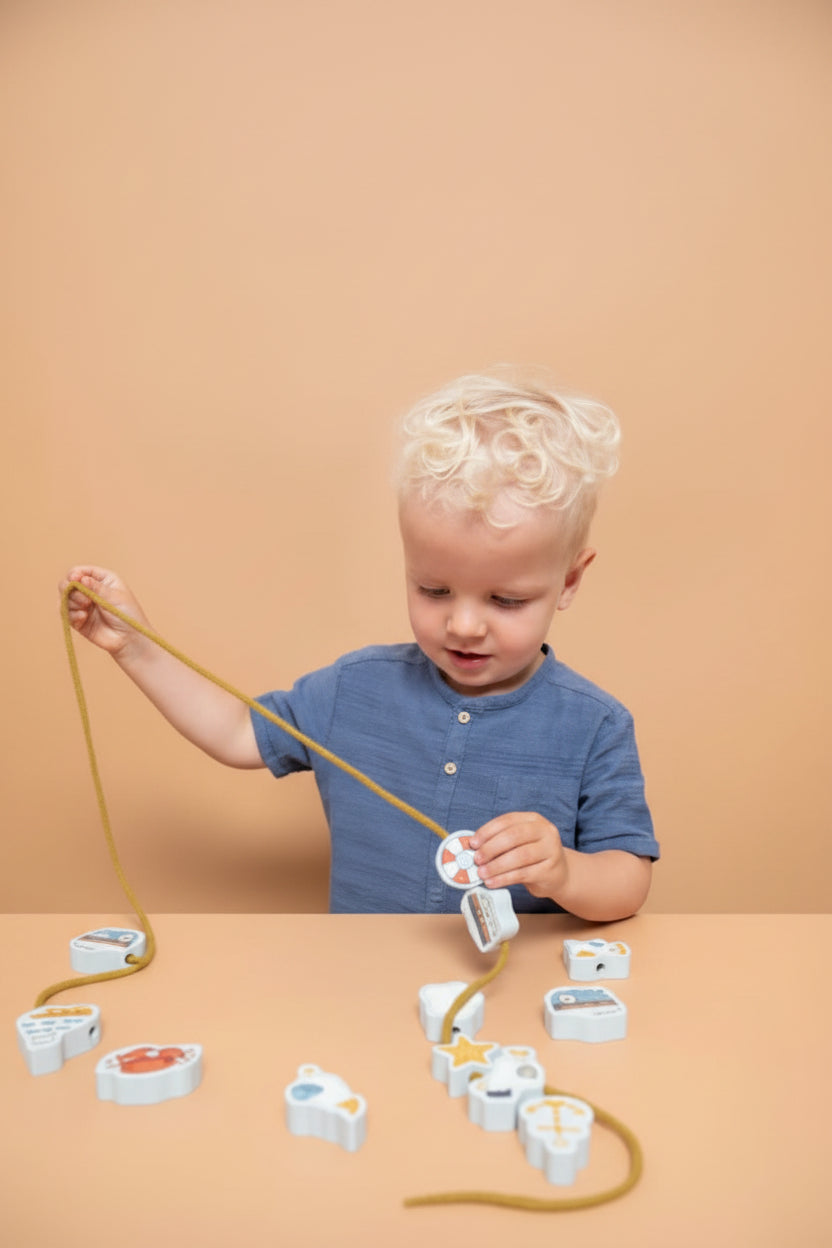 A young child with blonde hair wearing a blue shirt is sitting at a table playing with a beading set,  and reel while several small fish-shaped pieces are scattered on the table against a solid beige background.