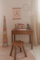 A cozy children's room corner featuring a light wood writing desk with a scalloped front, a matching flower-shaped wooden stool, and a stack of colorful numbered blocks on the floor. A pink coloring book, a pink patterned pencil case, and other school supplies are neatly arranged on the desk against a warm beige wall with a unicorn poster.
